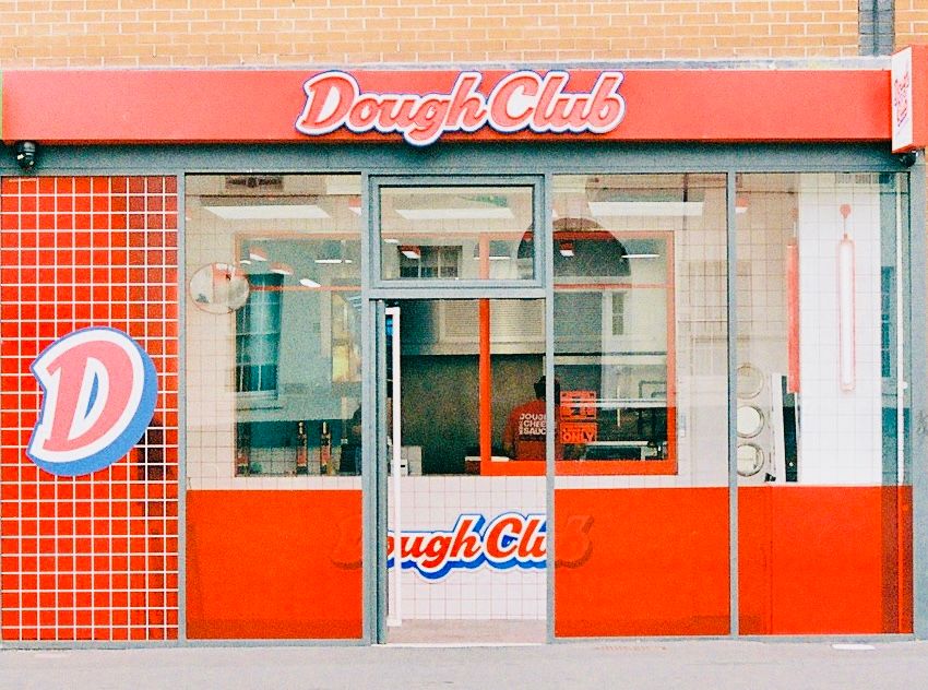 The Dough Club shopfront on Oxford Road in Manchester, a full-width glass frontage under a red fascia with the Dough Club sign, and the blue D logo on red tiling to the left of the door.